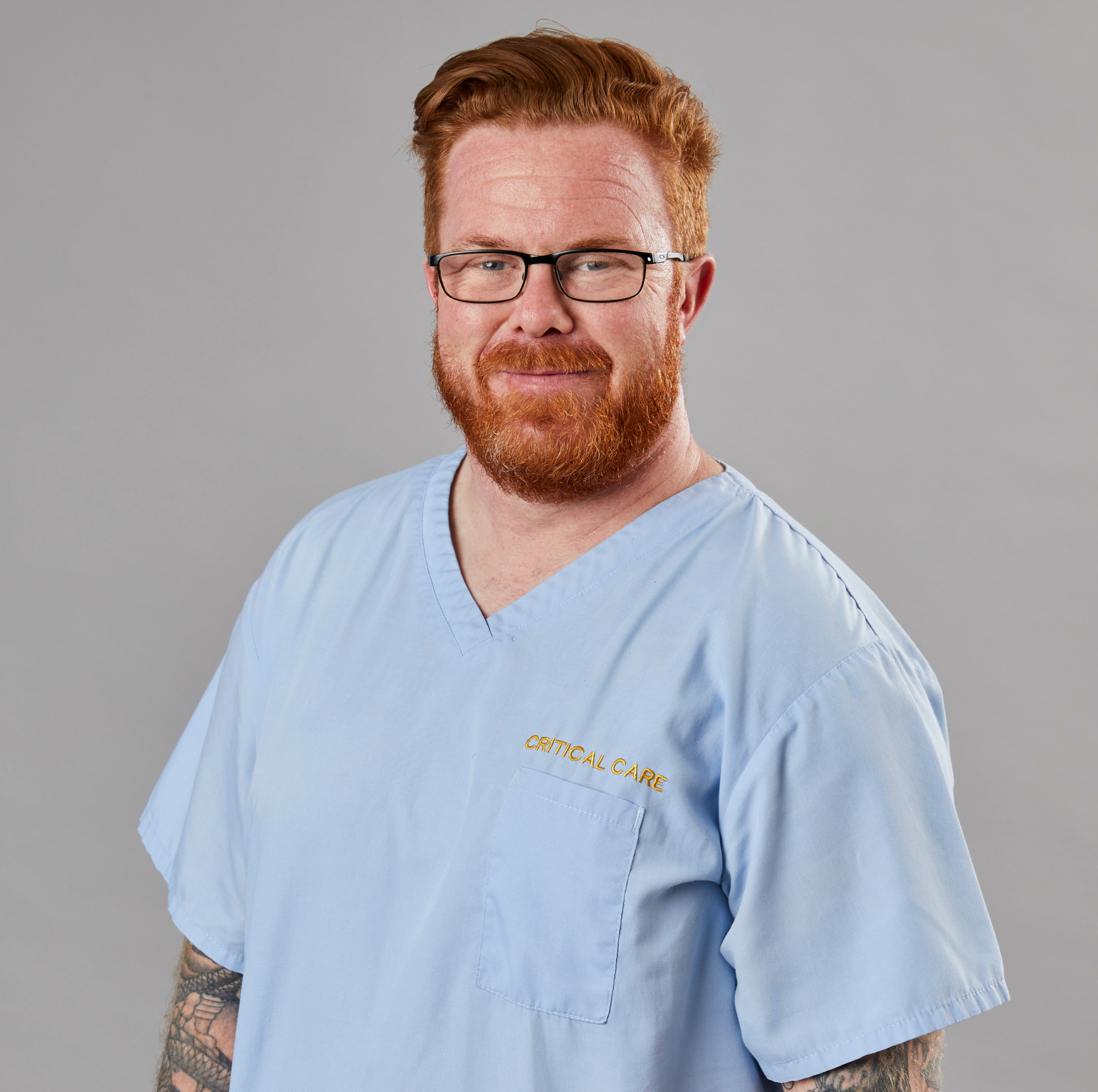 A man with a beard wearing glasses and a light blue scrubs labeled "critical care," smiling gently, with visible tattoo sleeves, standing against a grey background.