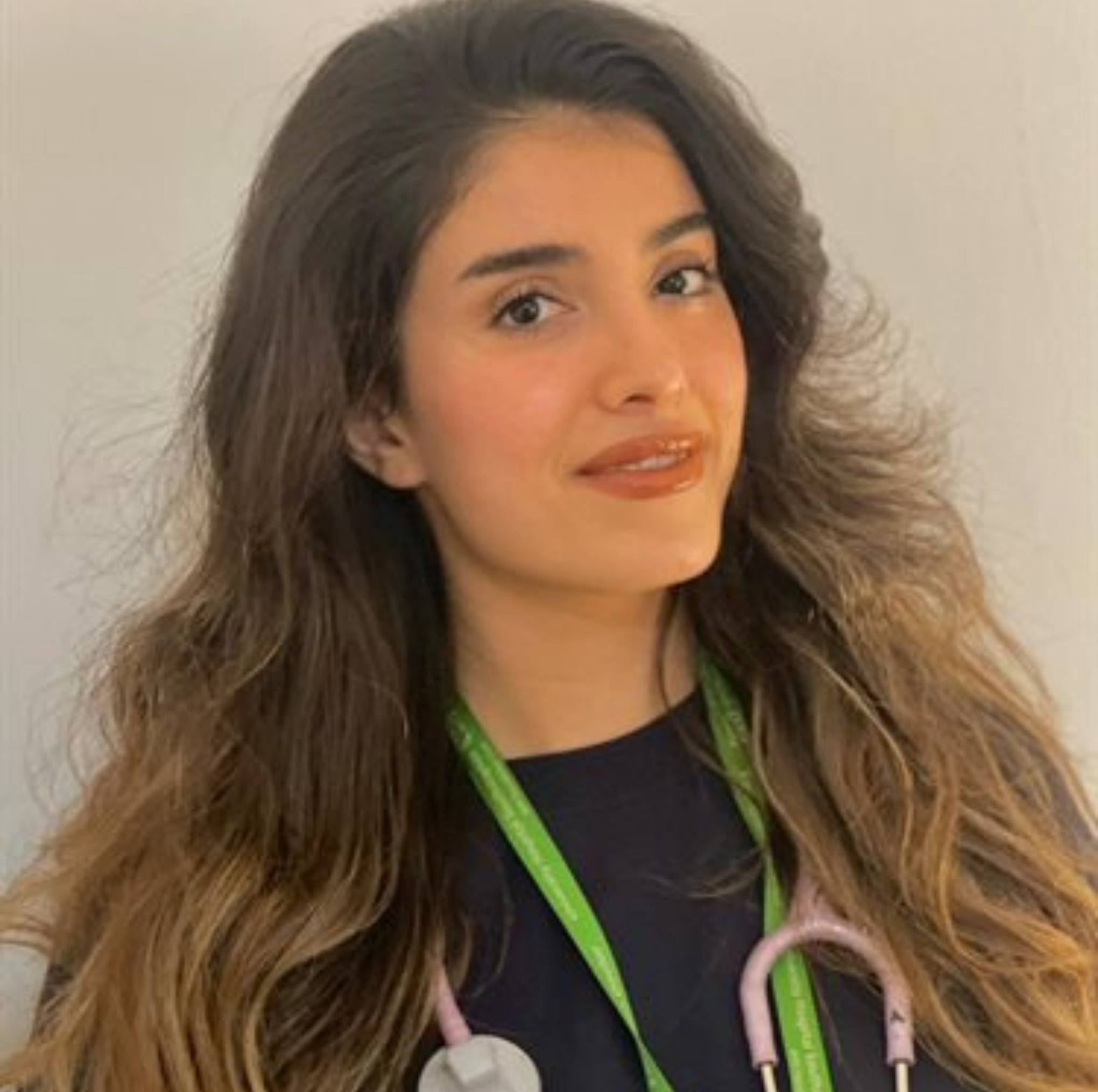 A young female healthcare professional with long, wavy brown hair wearing a stethoscope around her neck and a lanyard, smiling gently at the camera.