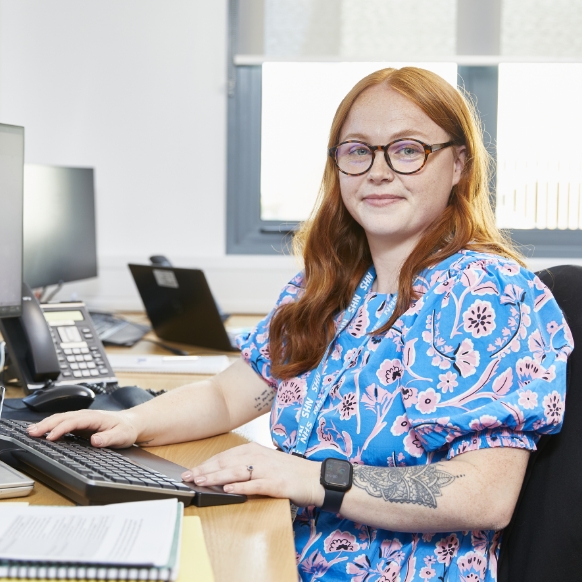 A woman with red hair and glasses, wearing a blue floral shirt, sits at a desk working on a computer in an office setting. she has a visible tattoo on her left arm.