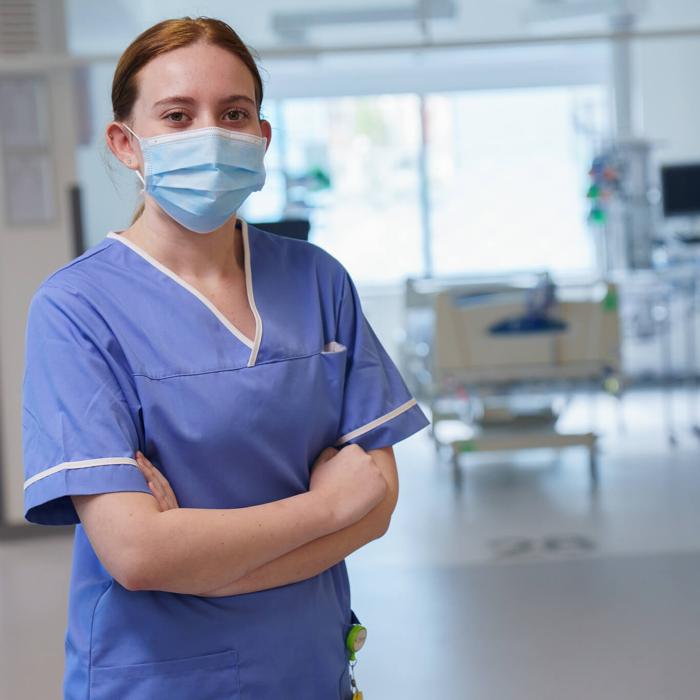 A female healthcare professional wearing a blue scrub and a surgical mask, standing with crossed arms in a hospital setting with medical equipment in the background.
