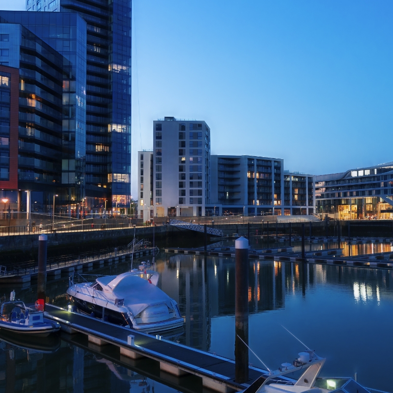 Twilight view of a tranquil marina with modern buildings and calm waters reflecting soft lights; boats docked along a wooden jetty.