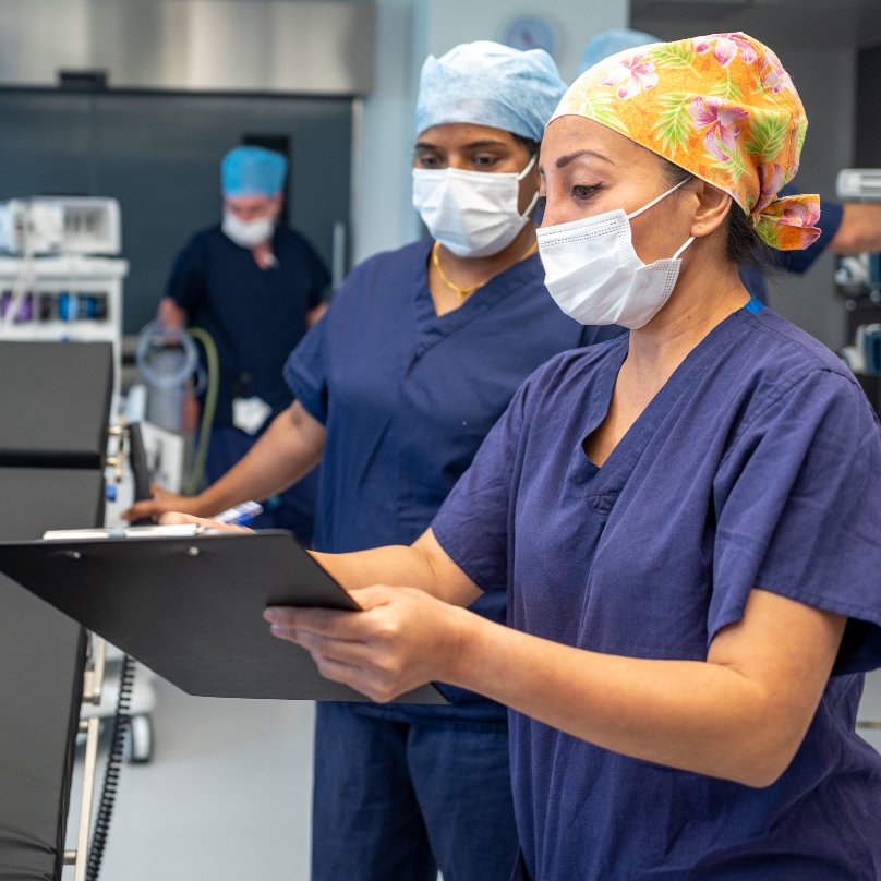 Two healthcare professionals in scrubs and surgical masks review a clipboard in a hospital setting with medical equipment in the background.
