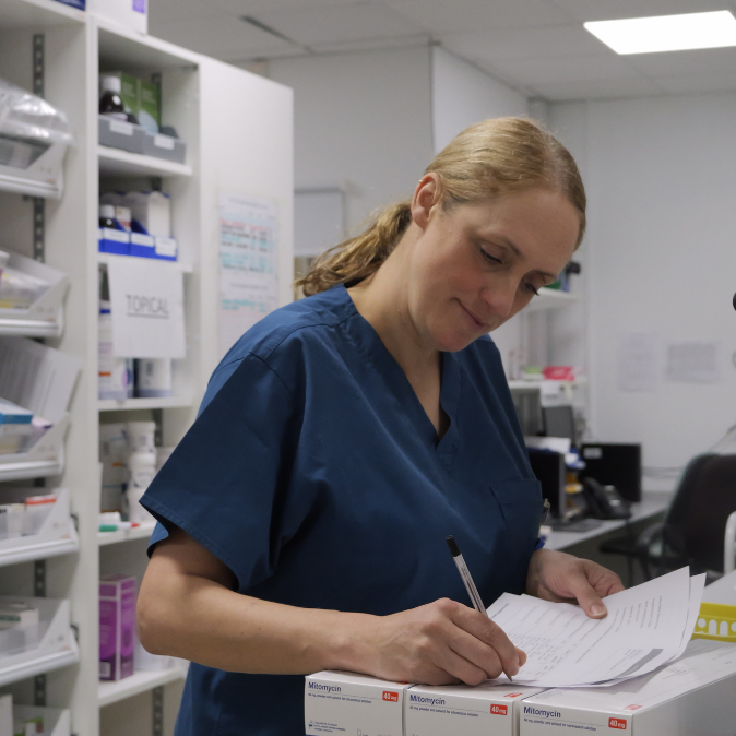 A female healthcare worker in blue scrubs is writing on a document, standing in a pharmacy with shelves stocked with medical supplies in the background.