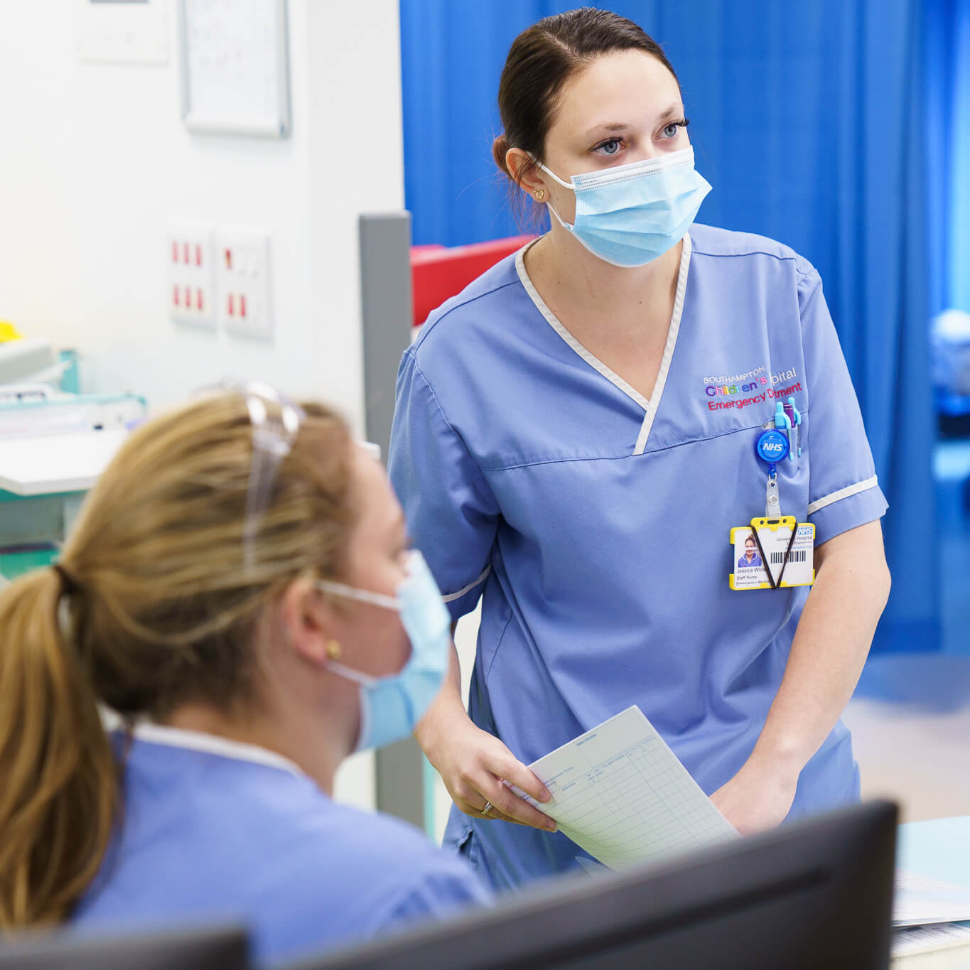 A female healthcare worker wearing a blue scrub and a mask discusses with another seated female colleague in a hospital setting. both are focused and attentive.