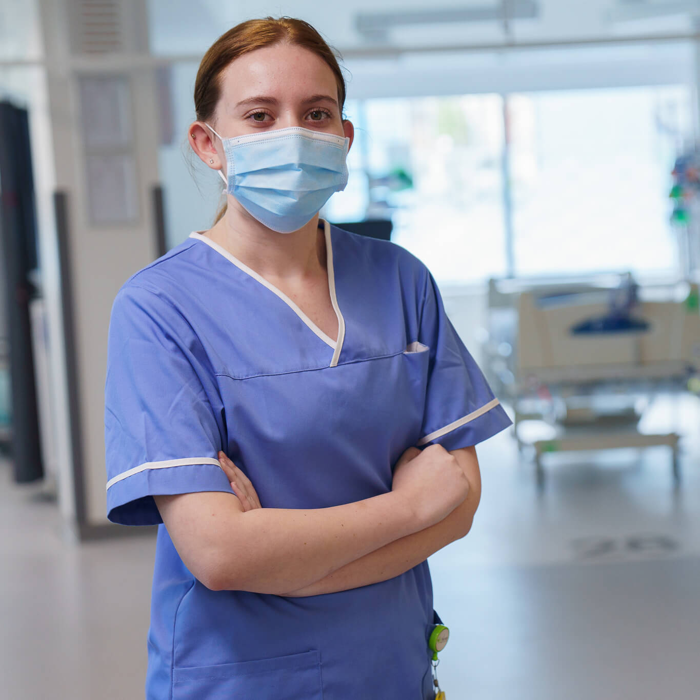 A female nurse wearing a blue scrub and a surgical mask stands with her arms crossed in a hospital corridor, looking directly at the camera.