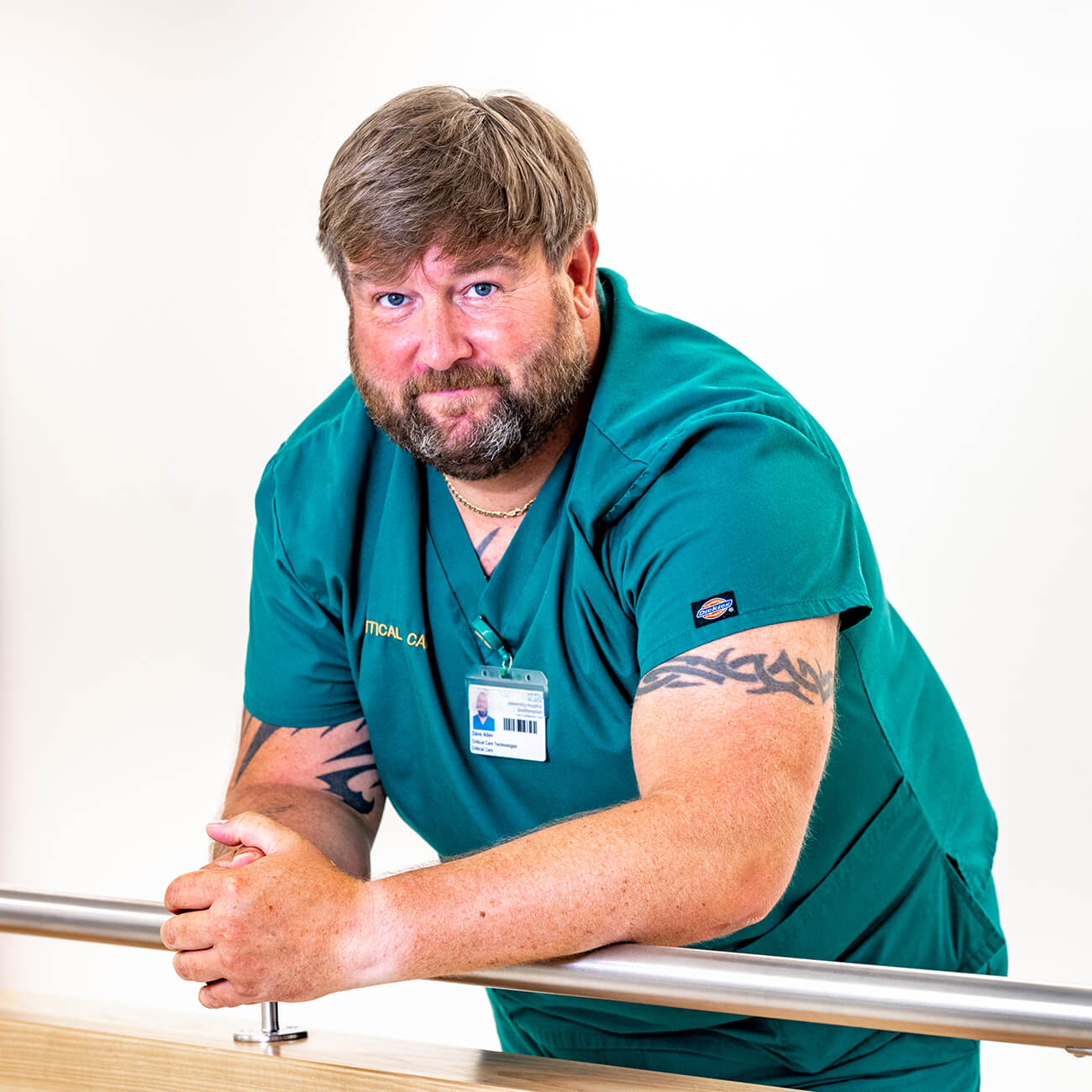 A male healthcare professional with a beard, wearing green scrubs and an id badge, leaning on a railing in a clinical setting and looking at the camera.