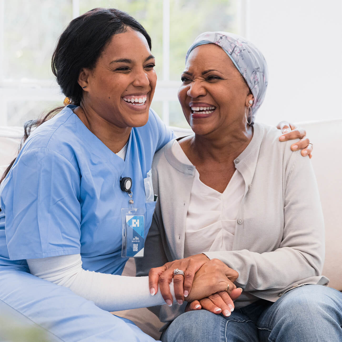A female nurse in blue scrubs shares a joyful moment with an older woman wearing a headscarf, both laughing together in a brightly lit room.