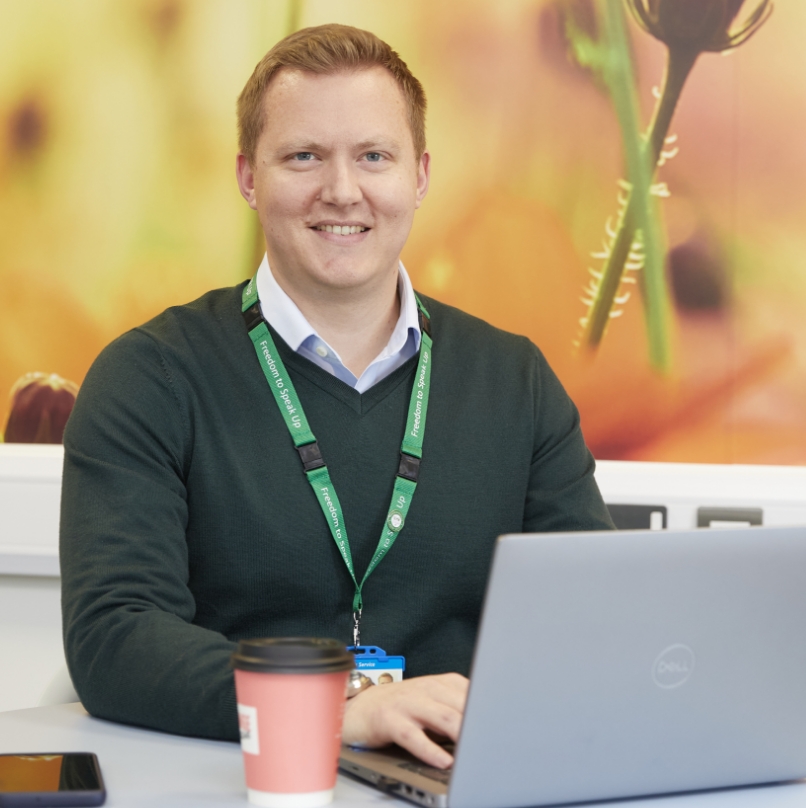 A smiling man in a green sweater and collared shirt sits at a desk, working on a laptop. he wears an id badge and there is a coffee cup on the desk, with a vibrant floral background.