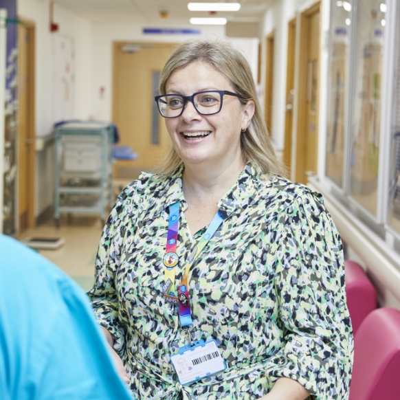 A cheerful female healthcare worker with glasses, wearing a patterned blouse and id badge, standing in a hospital corridor, engaging in conversation.