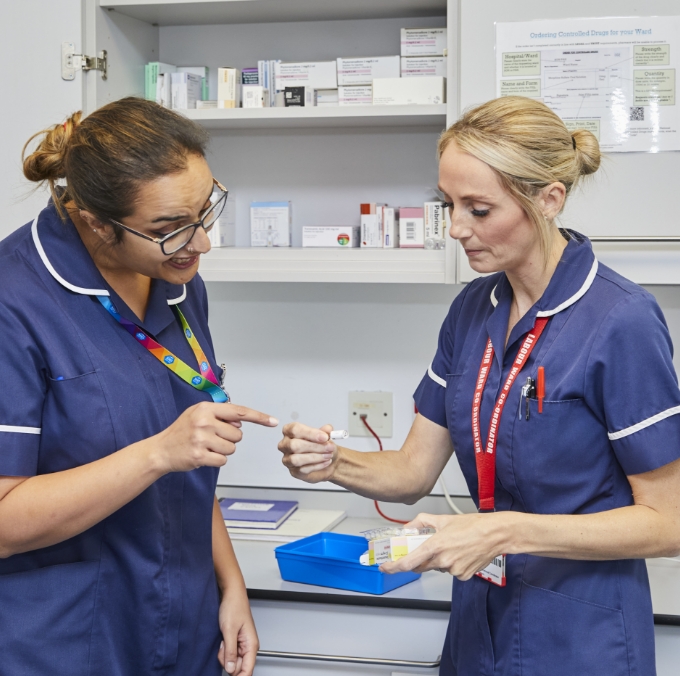 Two female nurses, one pointing at a syringe, discuss medications in a hospital's supply room with shelves of medical supplies in the background.