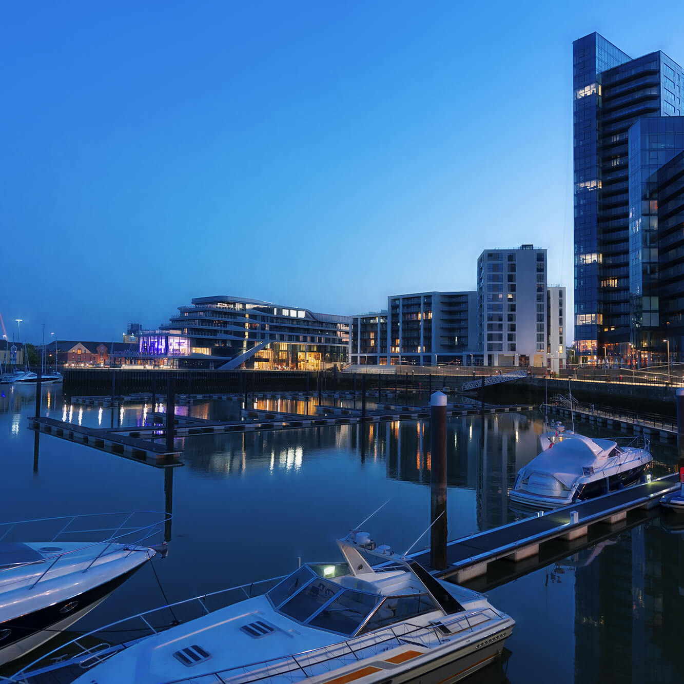 A serene twilight scene at a modern marina with sleek yachts docked along the water, surrounded by contemporary buildings under a clear blue sky.