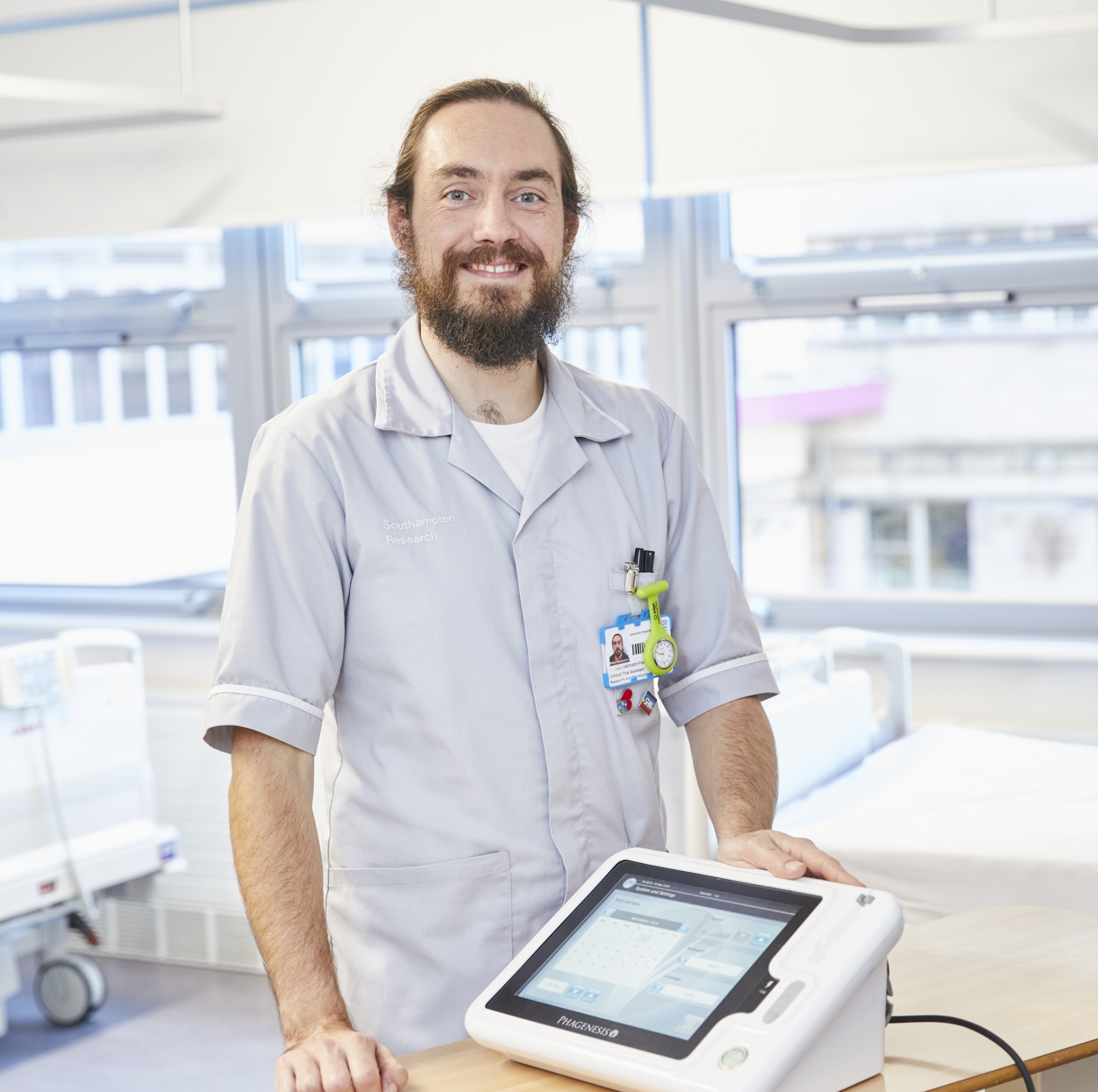 A smiling male healthcare worker with a beard, standing in a hospital room beside a medical device on a trolley. he wears a light grey uniform and an id badge.