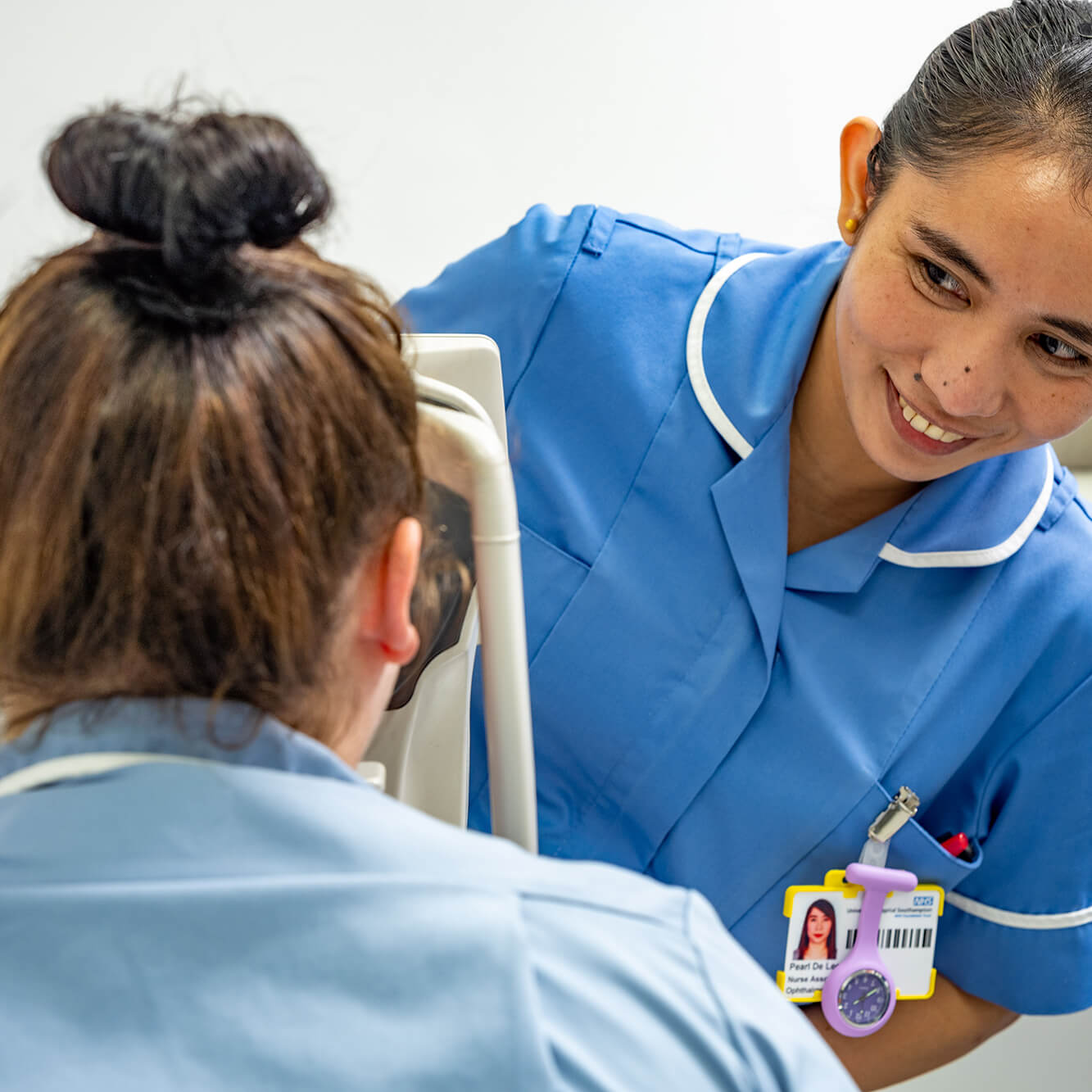 A smiling female nurse in a blue uniform assists a female patient seated in a medical chair, focusing on her care. the nurse wears an id badge.