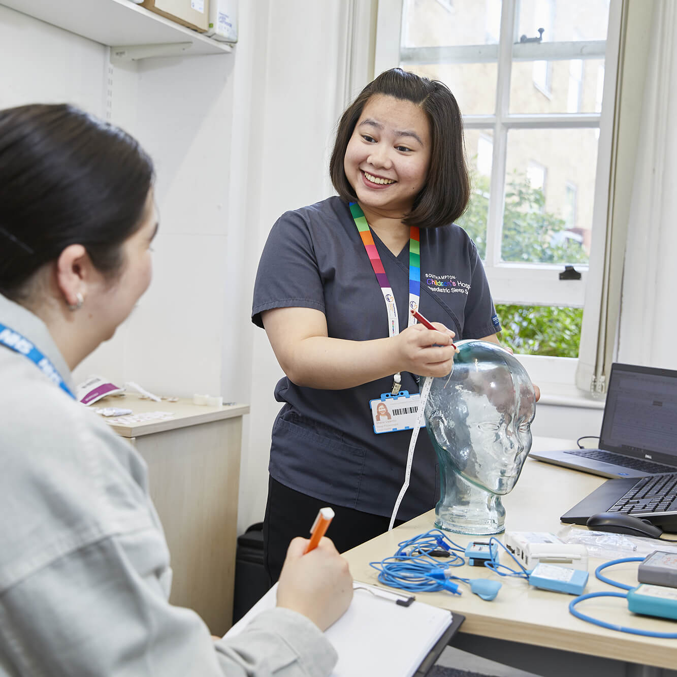 A smiling female healthcare instructor with a badge demonstrates medical equipment to a female student in a classroom, using a glass model of a head.