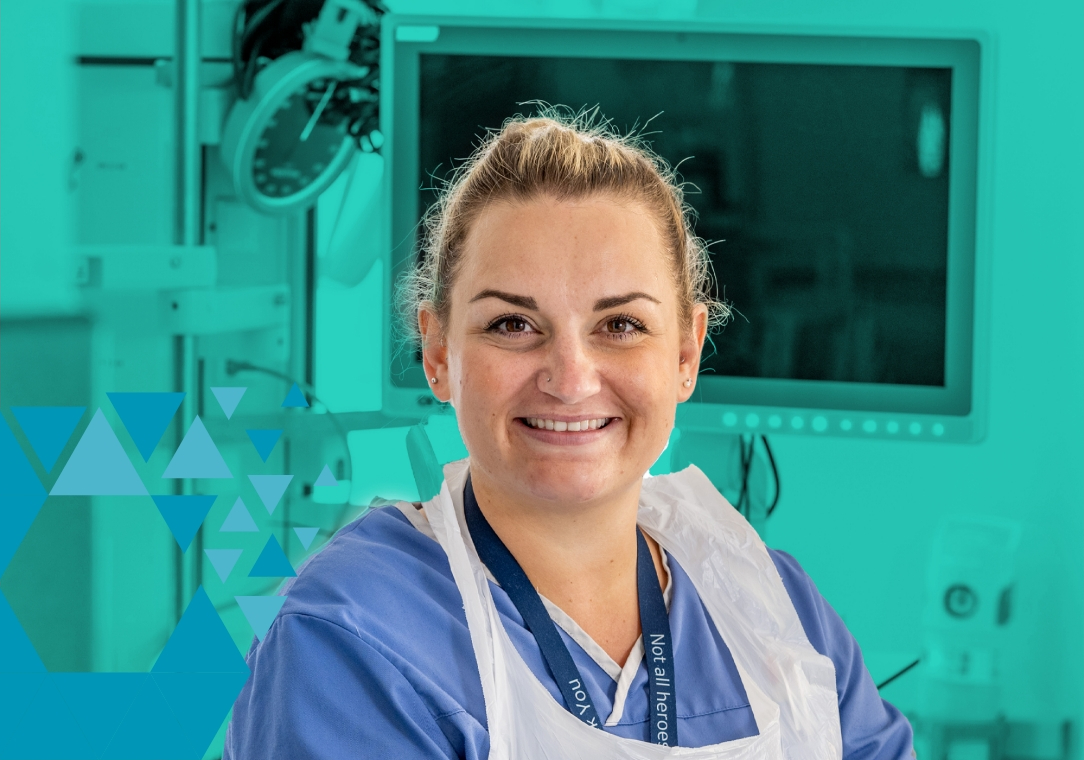 A cheerful female nurse in blue scrubs and a lead apron smiling in a hospital room with medical equipment in the background.