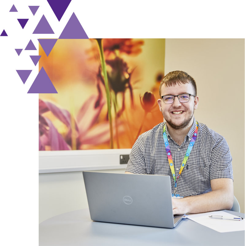 A smiling man wearing glasses and a plaid shirt sits at a desk with a laptop, against a background featuring a large floral photograph and abstract purple shapes.