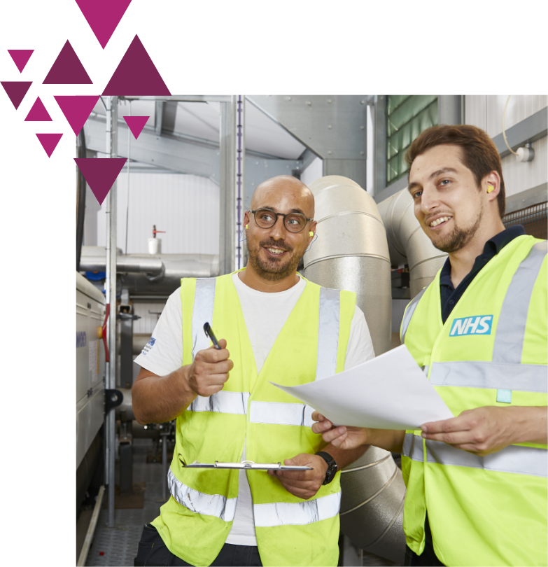 Two men in nhs high-visibility vests stand in an industrial setting, holding papers and discussing. one man is bald, smiling, and holding a pen, while the other, younger, listens intently.