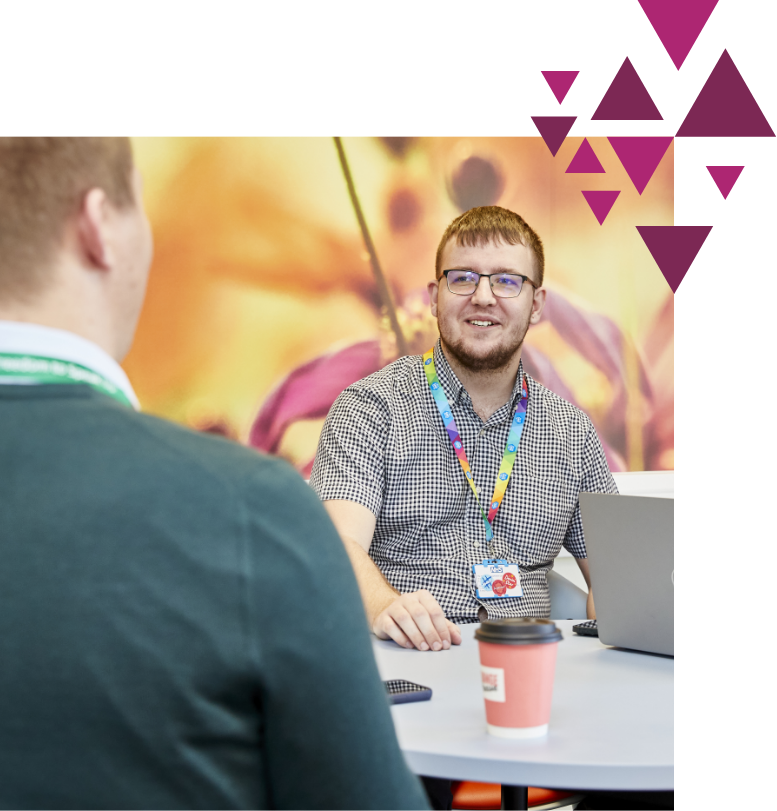 A man with glasses, wearing a plaid shirt and a lanyard, smiles while conversing with another man in a blurry foreground, sitting in a colorful office space.