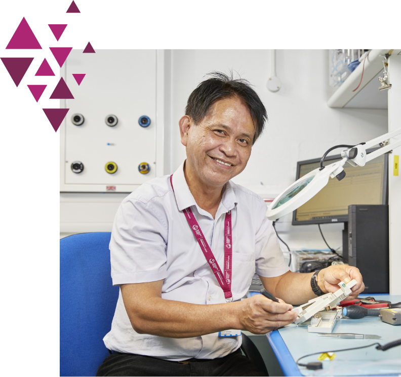 A smiling elderly asian man wearing a lanyard, sitting at a desk with electronic equipment, working on a component with a magnifying lamp in a brightly lit office.