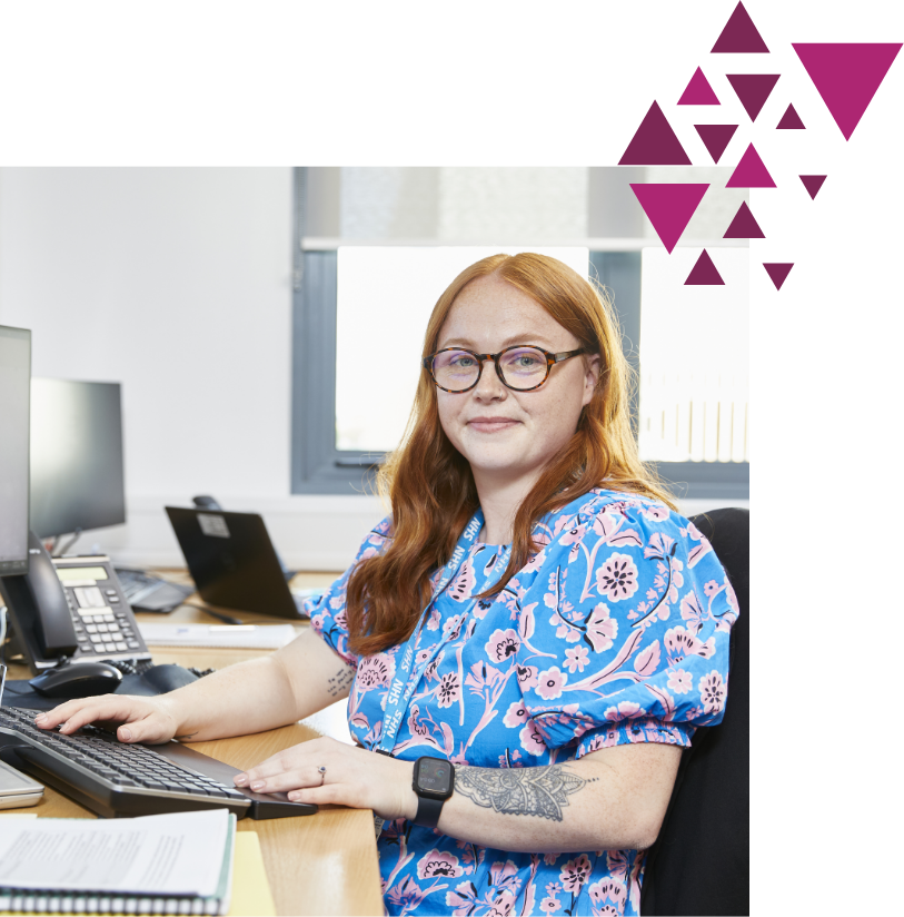 A woman with red hair and glasses, wearing a blue floral blouse, sits at a desk with computers, smiling at the camera, with pink abstract shapes in the corner.