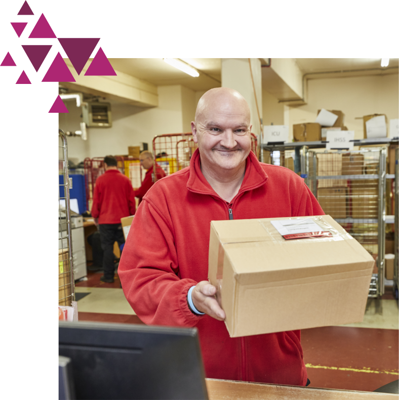 A smiling man in a red sweatshirt holding a cardboard box in a bustling warehouse environment, with coworkers active in the background.