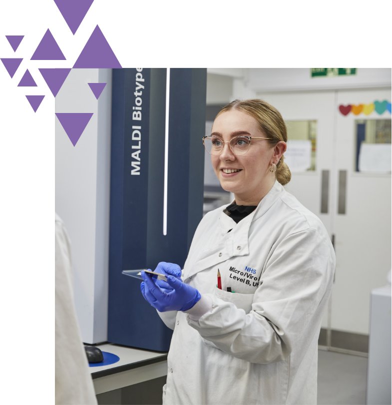 A female scientist in a lab coat, smiling and holding a smartphone, stands in a laboratory with maldi biotyper equipment visible in the background. decorative purple triangles accent the left side.