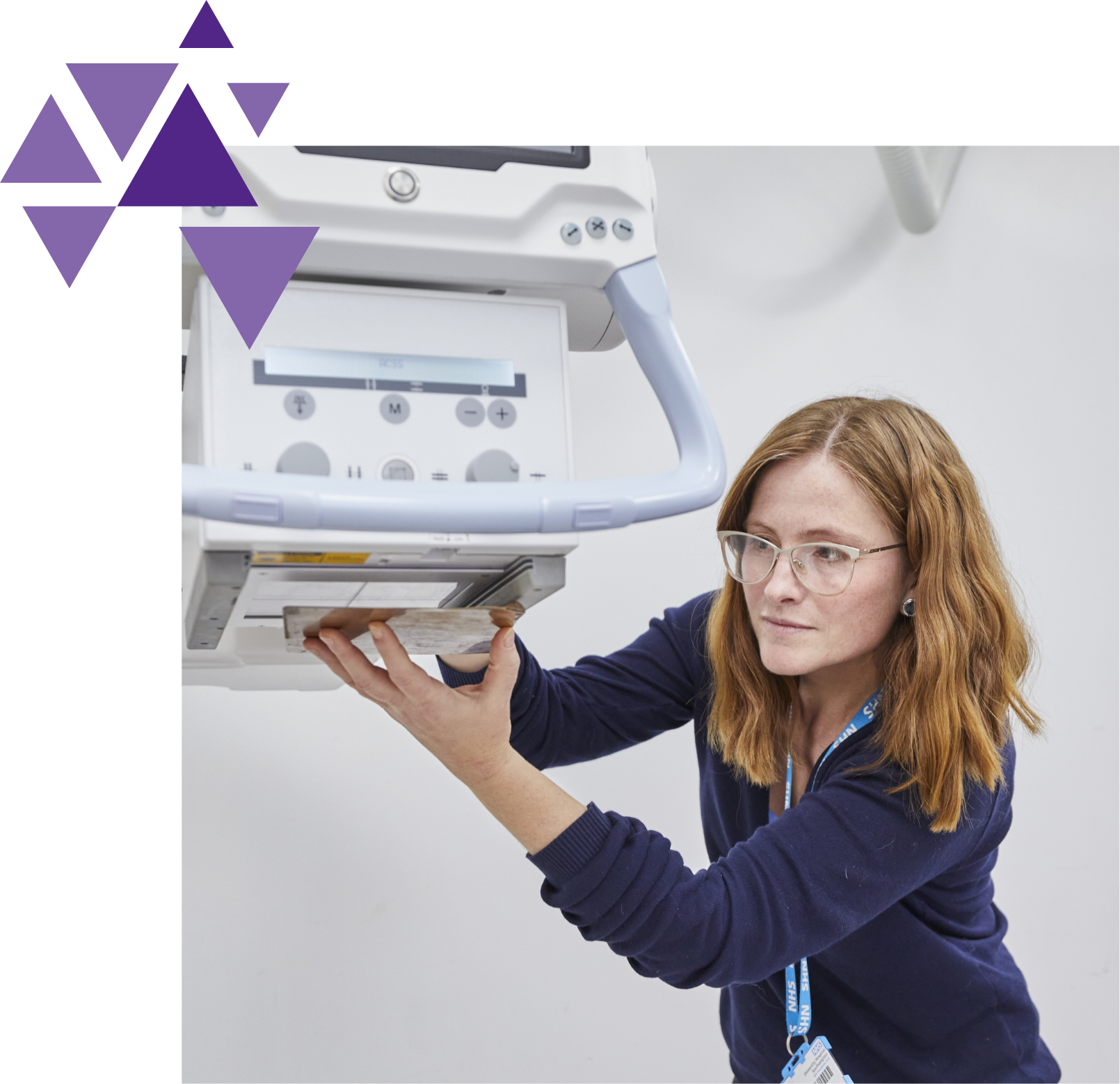 A female technician with glasses and a badge operates a large medical device, focusing intently on adjusting or examining a component.