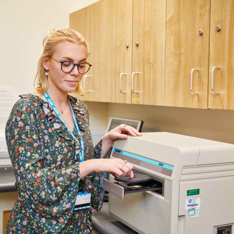 A woman with blonde hair, wearing glasses and a floral dress, uses a printer in an office setting, with wooden cabinets in the background.