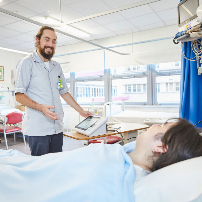 A male nurse standing next to a patient's bed in a hospital room, smiling and conversing with the patient who is lying down. there is medical equipment nearby.