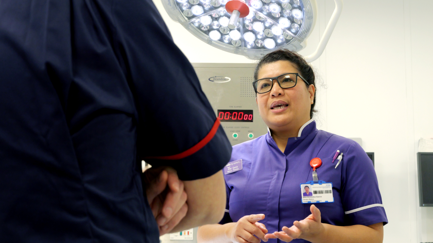 A female nurse in purple scrubs converses with a colleague under bright surgical lights in a hospital setting.