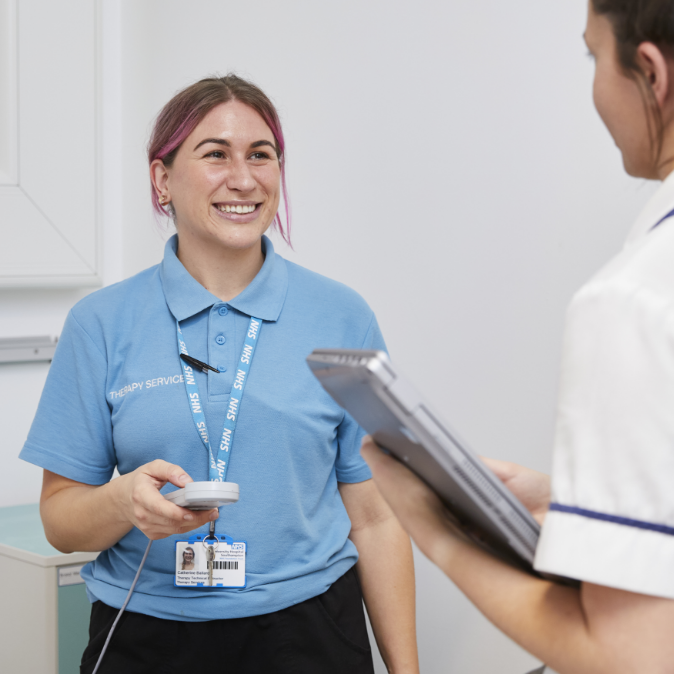 A female healthcare worker in a blue polo shirt smiling at a colleague holding a clipboard, both standing in a clinical setting. the woman has pink hair and a badge reading "therapy services.