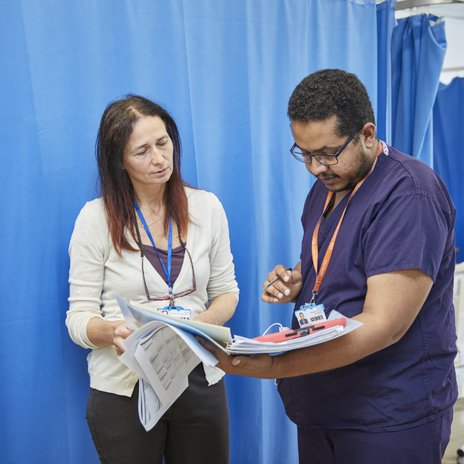 Two healthcare professionals, a man and a woman, wearing id badges, discuss a document in a medical facility with blue curtains in the background.