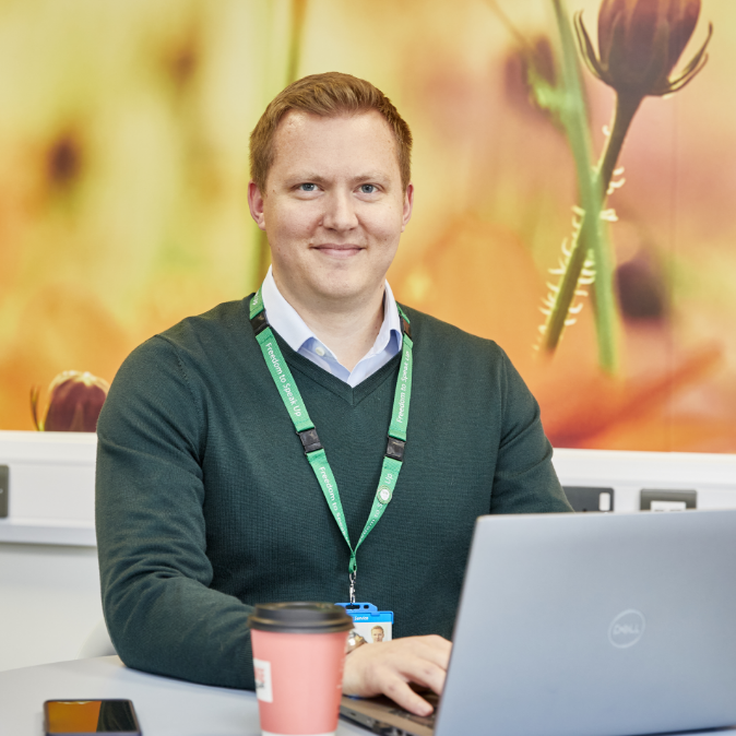 A man in a green sweater and blue shirt sits at a desk, working on a laptop, with a coffee cup next to him, in an office with floral wallpaper background.