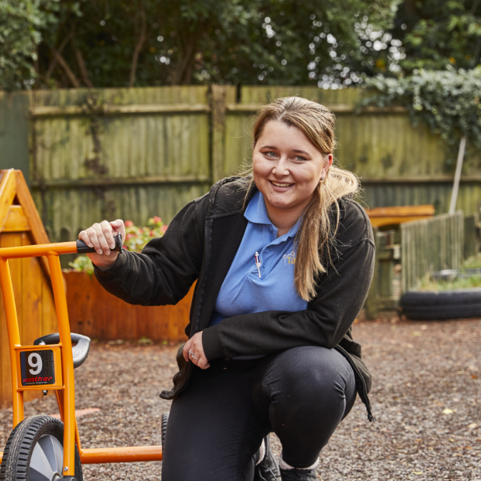 A woman in a blue jacket and black pants leans on a bright orange go-kart, smiling at the camera, with a wooden fence and greenery in the background.