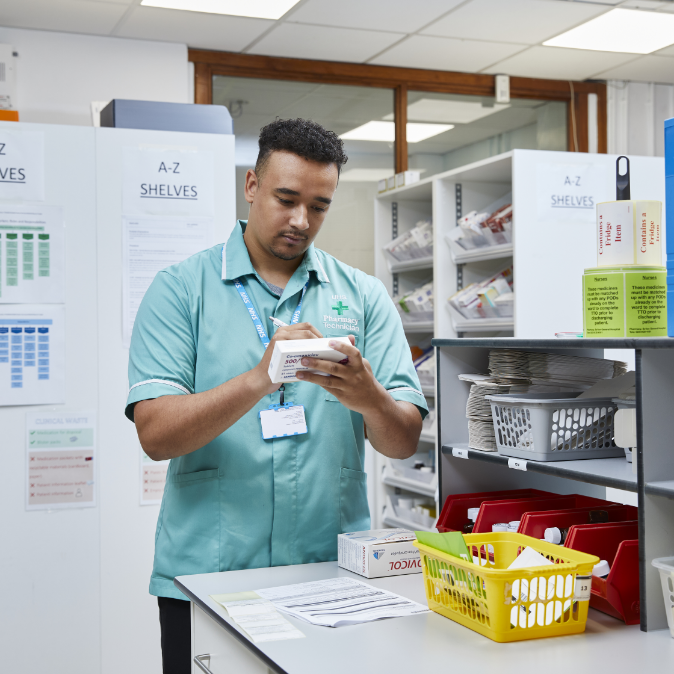 A male pharmacy worker in a green polo shirt checks inventory with a clipboard in a storeroom filled with shelves of medical supplies and boxes.