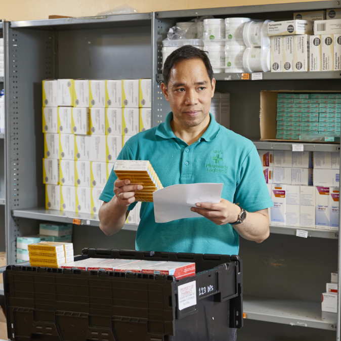 A male pharmacist in a teal uniform scans medication packages in a well-organized pharmacy stock room.