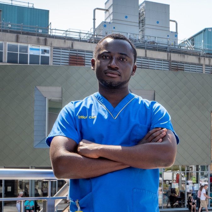 A confident male healthcare worker in blue scrubs stands with arms crossed in front of a hospital building. he is looking directly at the camera.
